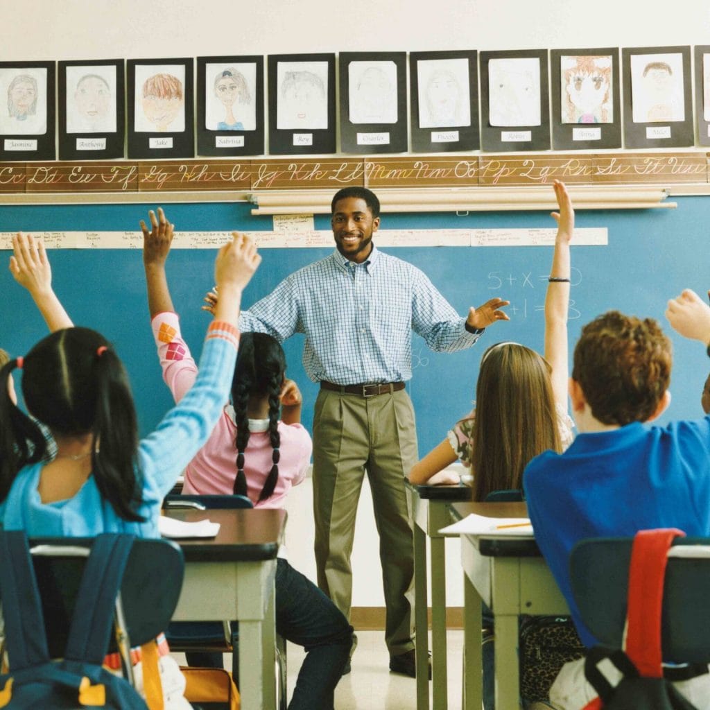 Teacher leading a class who are all raising their hands to answer his question