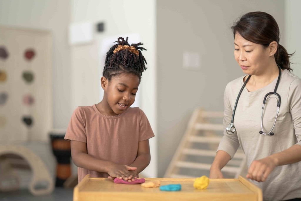 A student kneels down in front of a table with her therapist as she plays with some colorful modeling clay.